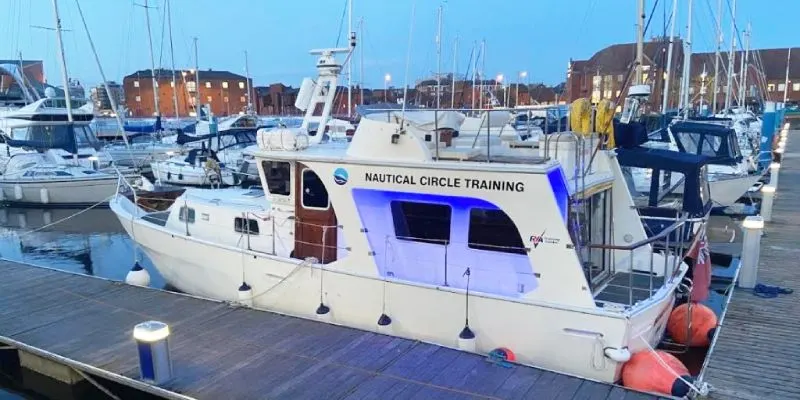 Moored training vessel with blue branding alongside other sailboats in a marina with brick buildings beyond.