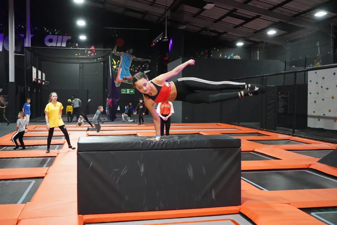 Woman performing aerial flip over foam obstacle on orange and black trampoline court with children and staff watching.