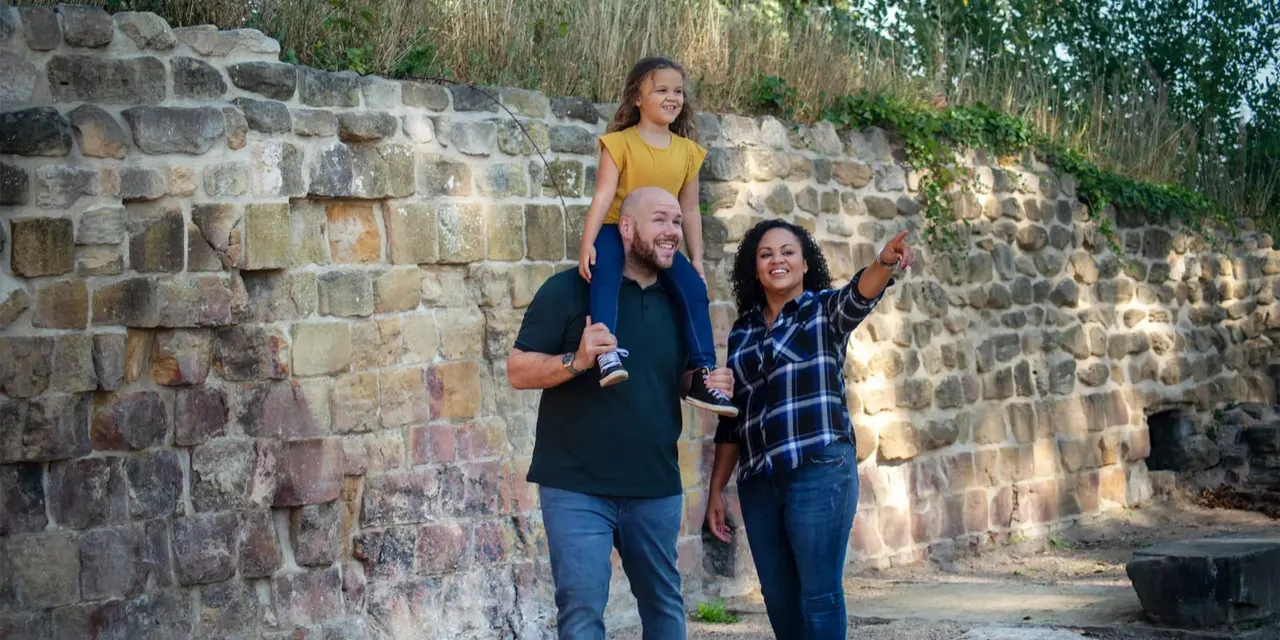 A man carrying a young girl on his shoulders walks with a woman past a historic stone wall, all smiling.