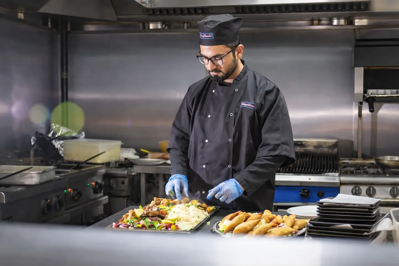 Chef preparing food on a counter in a commercial kitchen, with plated dishes and cooking equipment visible.