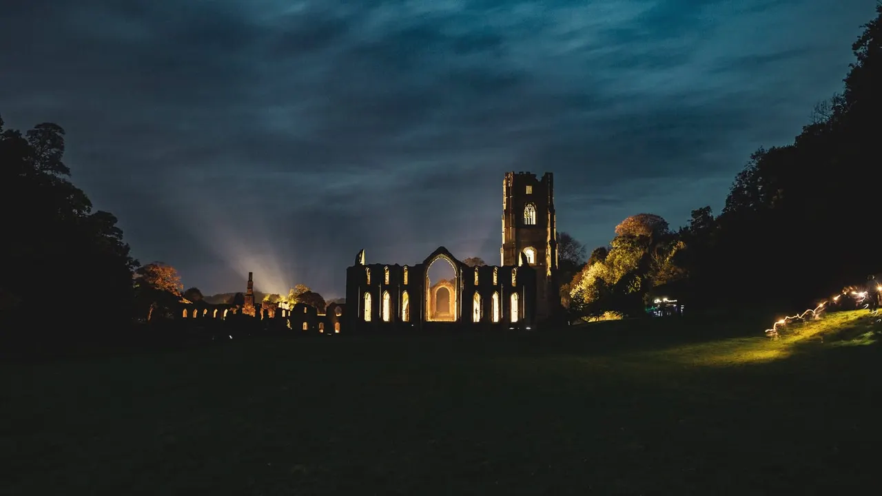 Illuminated ruins of a Gothic abbey at dusk, surrounded by trees and parkland under a stormy sky.