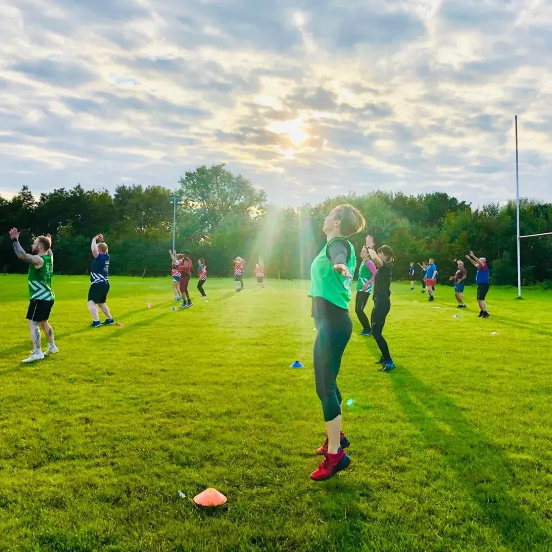 Large group of people exercising on a grass field at sunset, with training cones and a volleyball net visible.