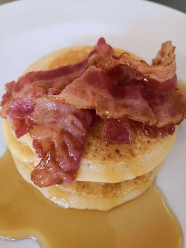 Stacked pancakes topped with crispy bacon strips, served on a white plate with maple syrup.