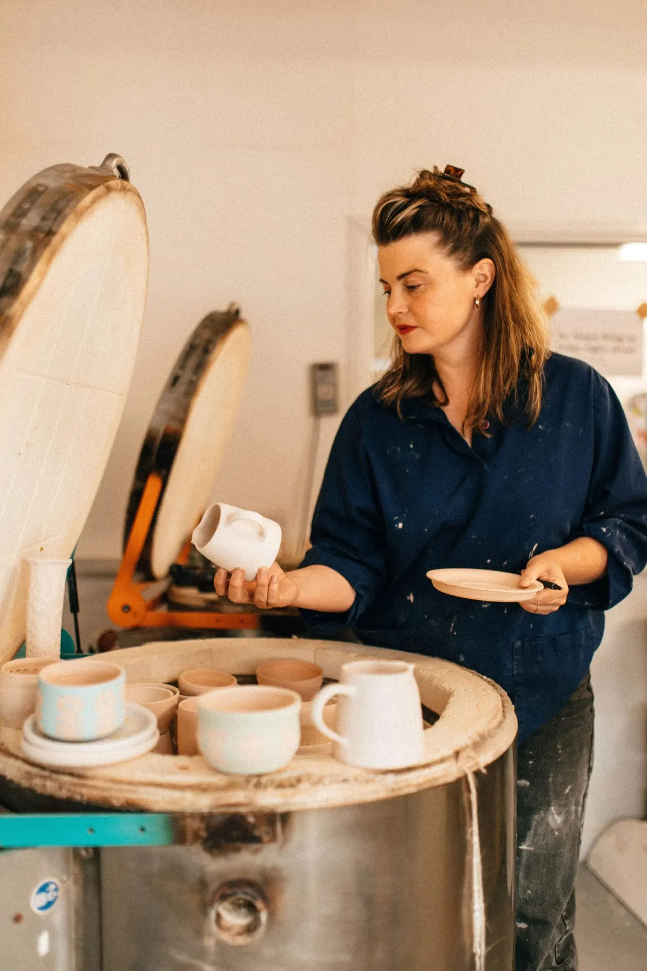 Woman examining a ceramic plate in a pottery studio, with a pottery wheel and finished white ceramic pieces displayed on a wooden table.