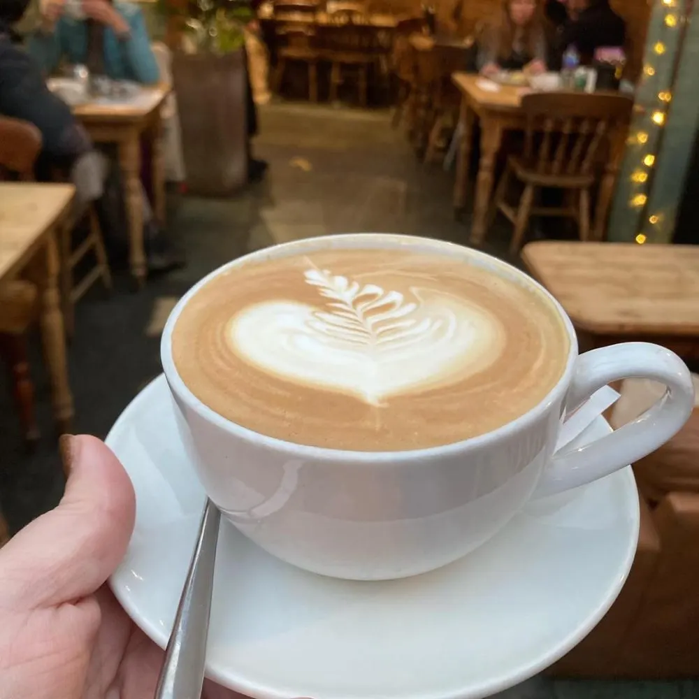 Hand holding a white cup of latte with latte art in a busy café interior with wooden chairs and diners.