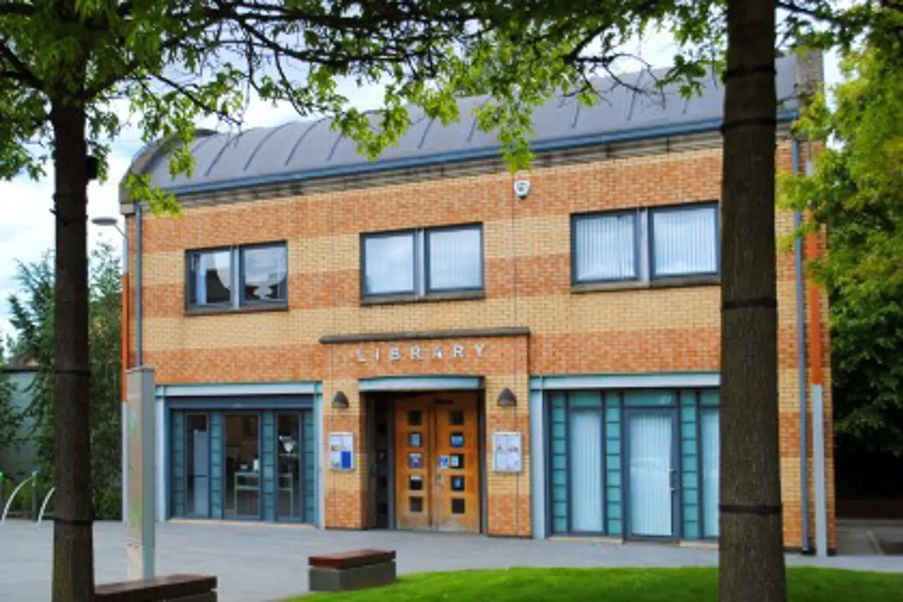 Modern brick library building with blue-trimmed entrance, viewed through trees in front courtyard.