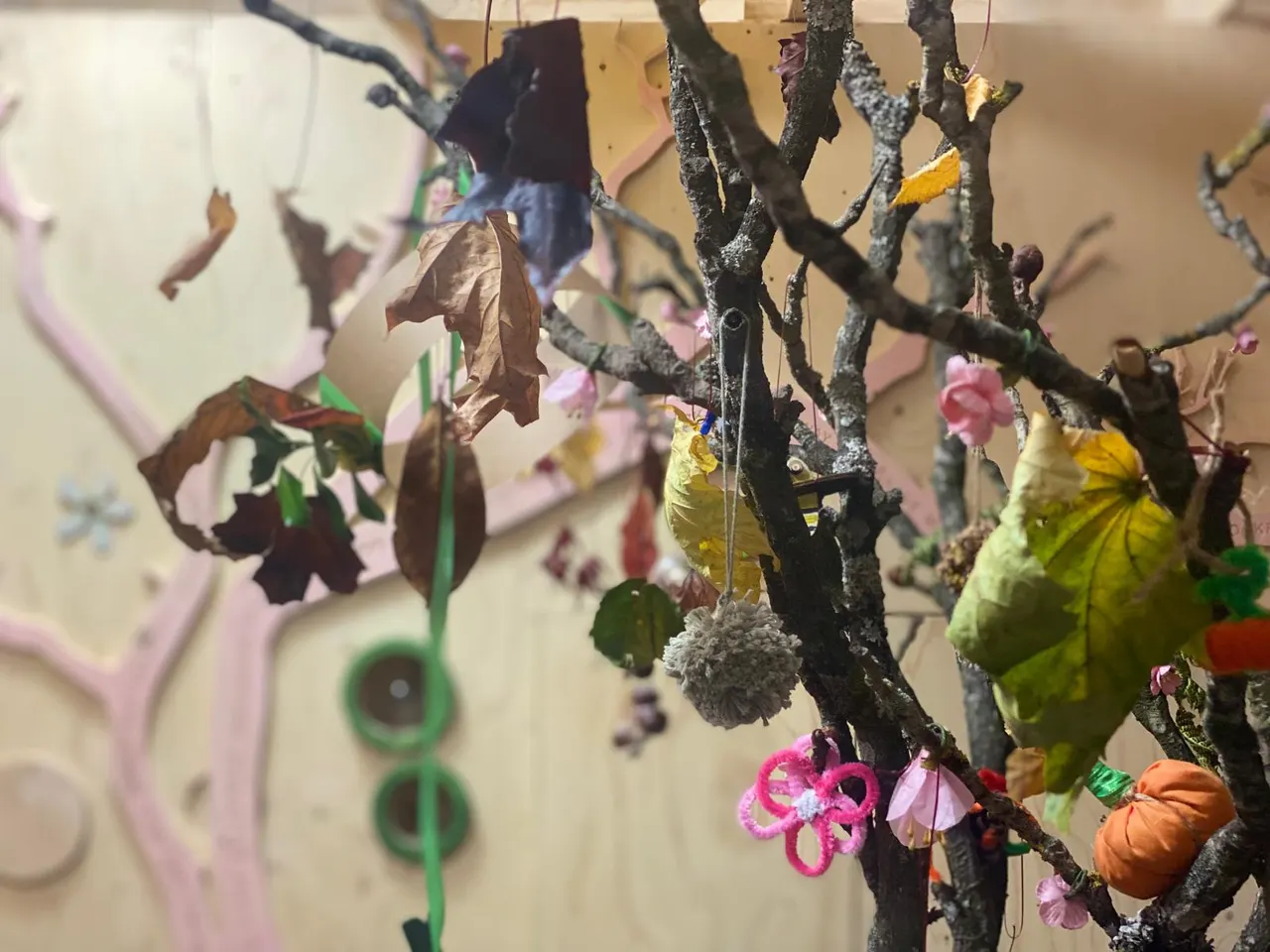 Bare branches decorated with colourful handmade ornaments including flowers, leaves and spheres hanging indoors.