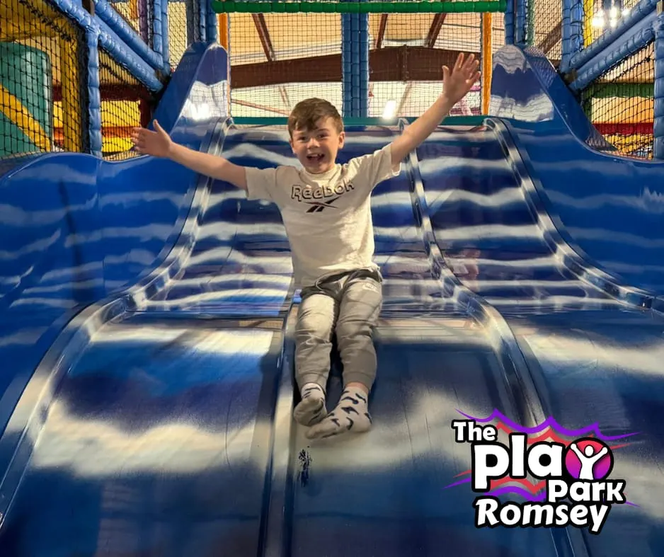 Young child sliding down a blue inflatable slide with arms raised, smiling.
