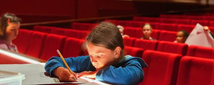 Child in blue shirt drawing or writing at a table in an empty theatre auditorium with red seats.
