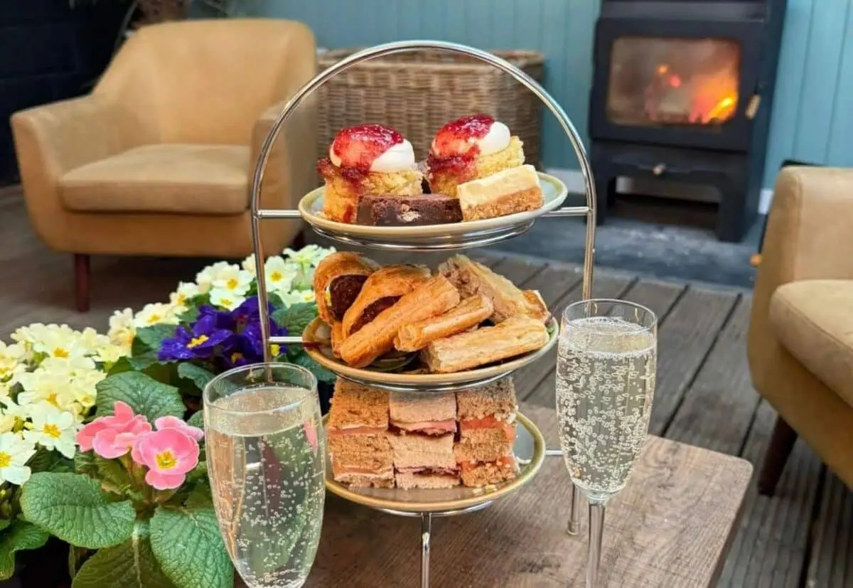 Three-tiered stand with pastries, cakes and sandwiches on a table by flowers and sparkling water, with fireplace and chairs behind.