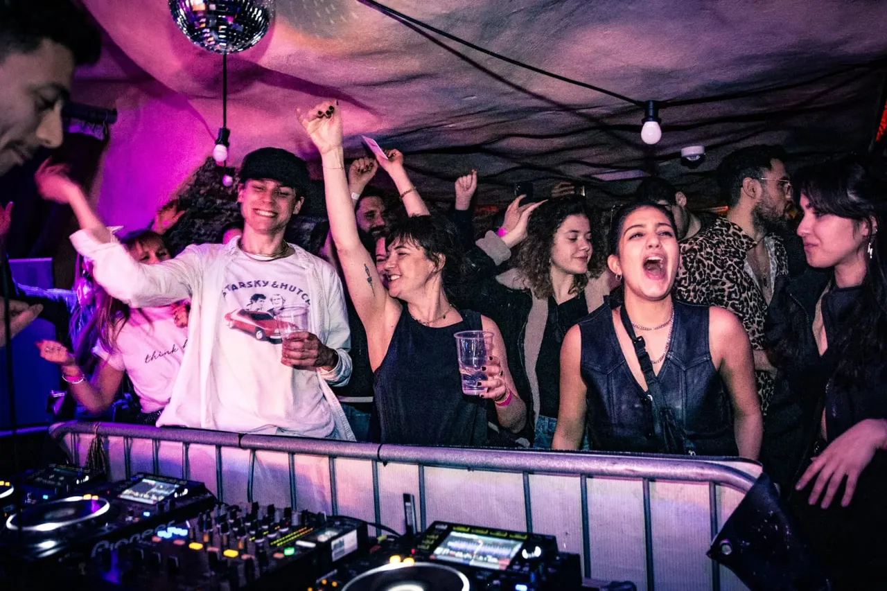 Crowd of people dancing and celebrating at a nightclub with purple lighting, disco ball, and DJ booth.