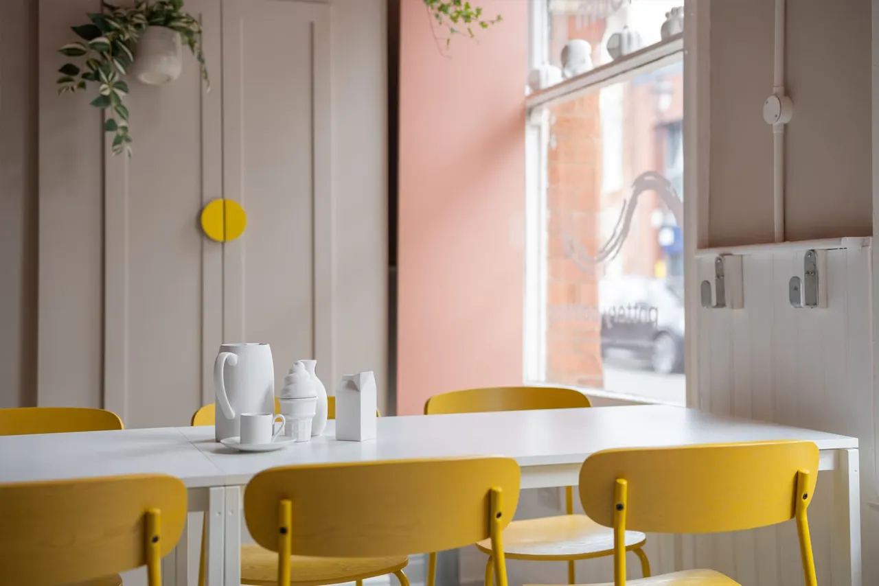 Modern café interior with yellow chairs at white tables, white crockery and hanging plants, overlooking a street.