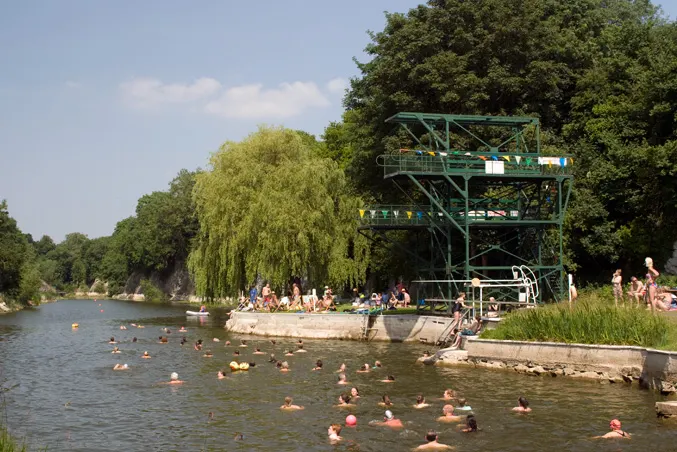 Swimmers and paddlers in a lake with a green diving platform and weeping willow trees lining the bank.