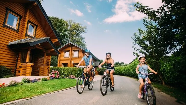 Three children cycling on a driveway past log cabin buildings on a sunny day.