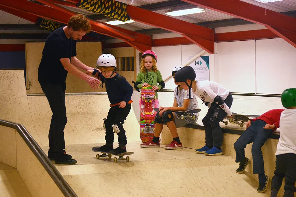 An instructor supervises young children learning to skateboard indoors, some wearing protective helmets and gear.