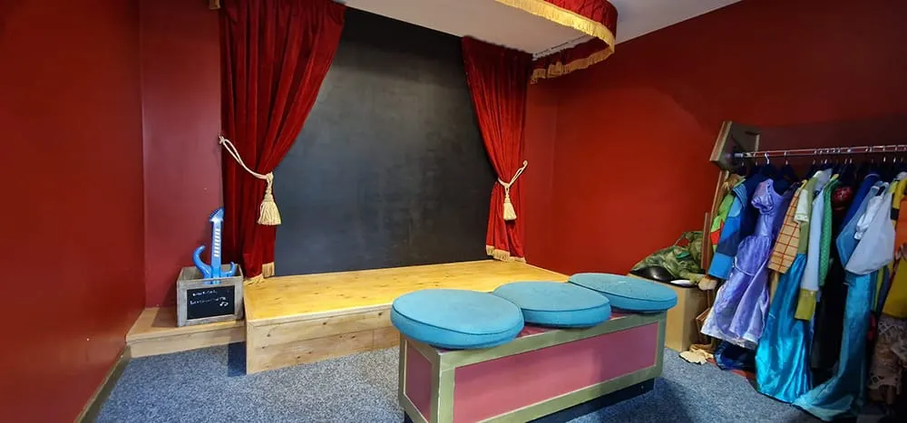A children's performance space with a wooden stage, red curtains, blue cushioned seating, and costumes hanging on the right wall.
