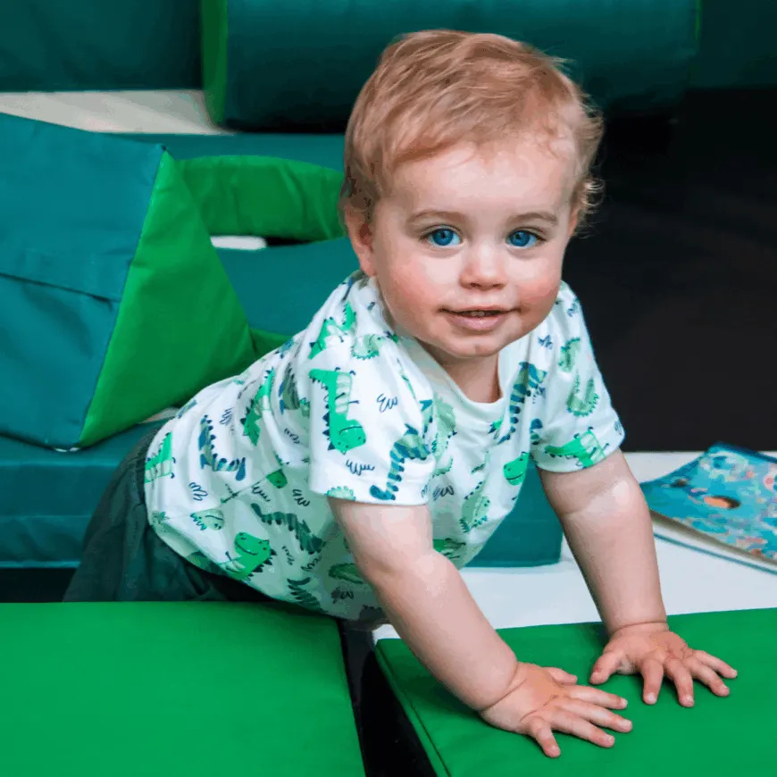 Toddler in patterned shirt crawling on green play structure with soft seating in background.