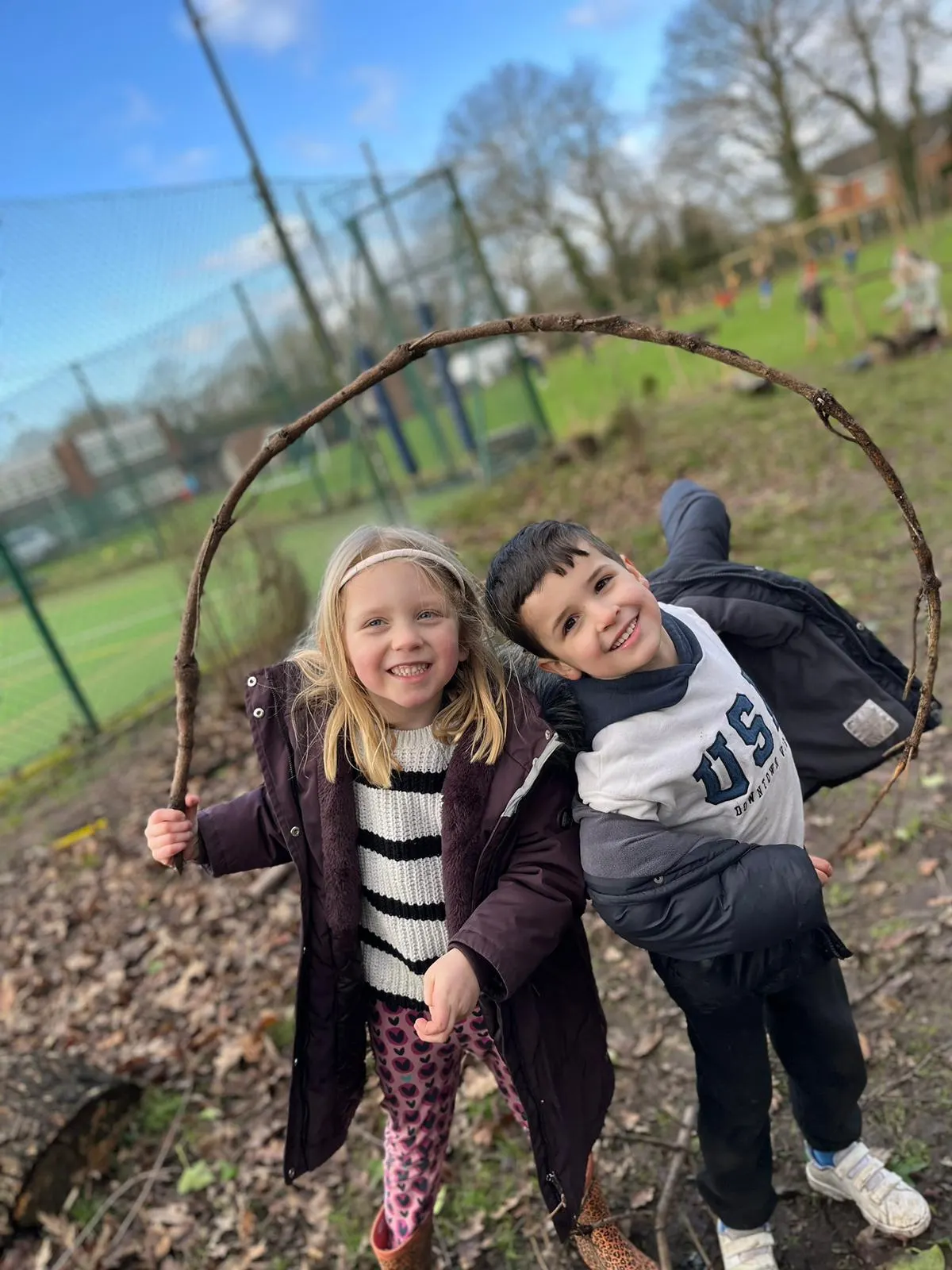 Two young children holding a curved stick together, smiling at a farm or outdoor activity venue.
