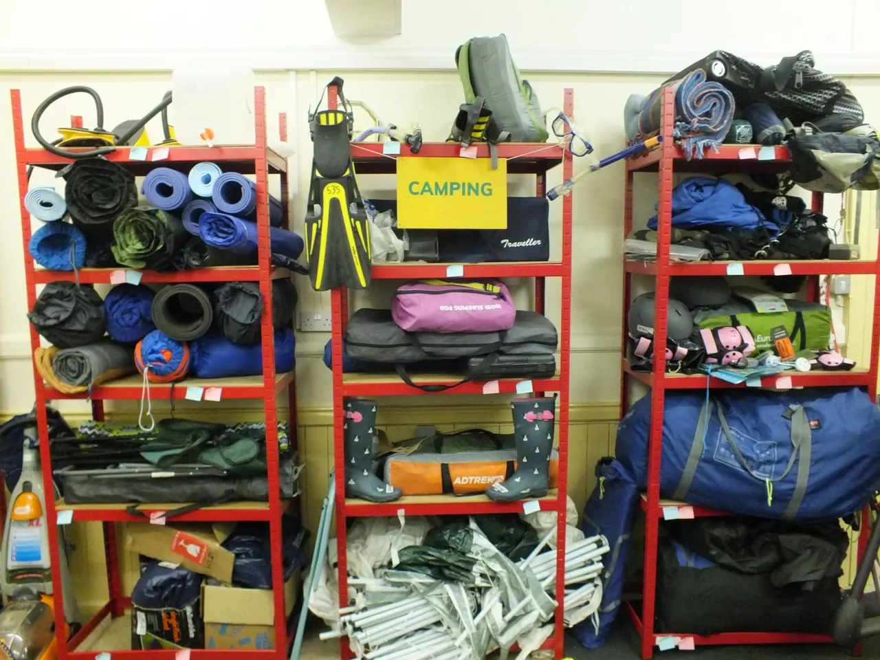 Red metal shelving units stacked with camping and outdoor equipment including sleeping bags, mats, fins, ropes and various gear available to borrow.