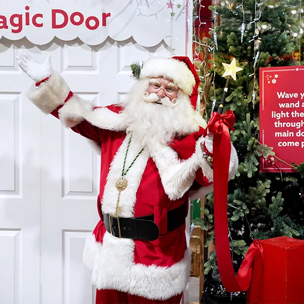 Person in Father Christmas costume waving at a white door decorated with festive garland and red bows.
