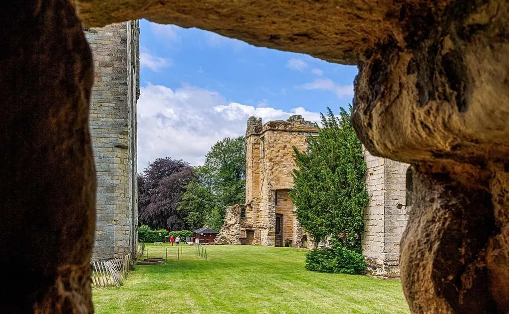 View through a stone archway of ruined castle walls with ivy and green grounds, framed by weathered masonry.