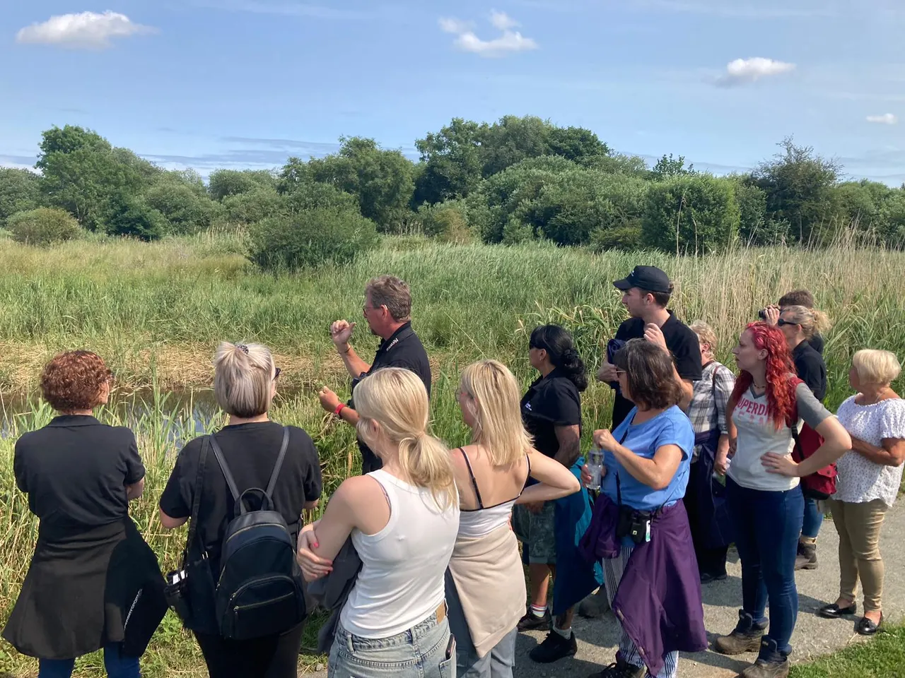 A group of visitors gathered on a path listening to a guide give an outdoor talk beside a reedbed and woodland under blue sky.