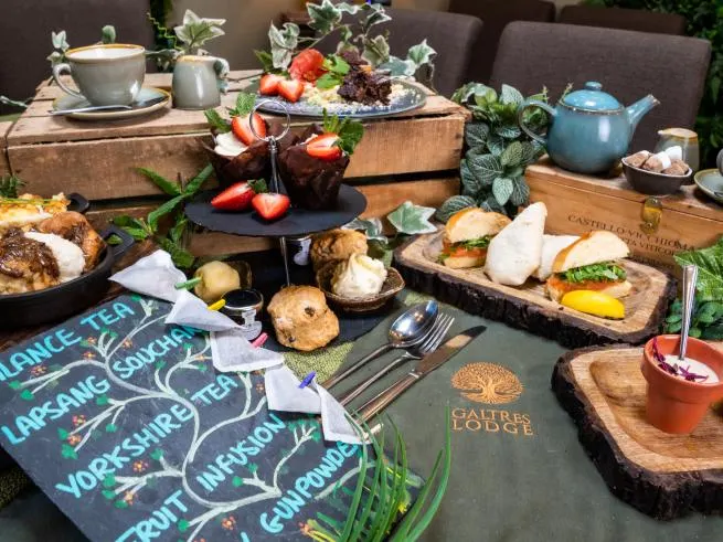 An elegantly styled afternoon tea spread with sandwiches, pastries, fresh berries and teapots arranged on wooden boards amongst potted plants and a slate menu board.