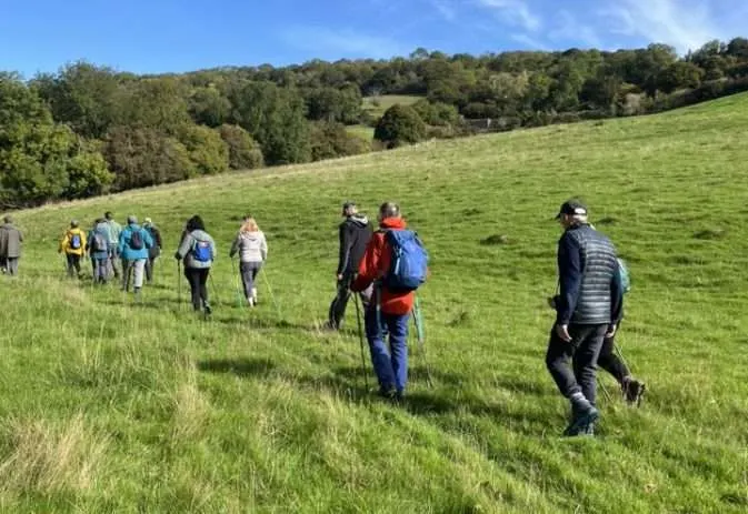 A group of people walking across a green hillside with woodland in the background under blue sky.