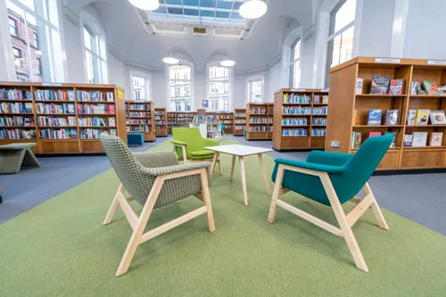 Library reading area with green and teal modern chairs around a white table on a lime-green carpet, surrounded by wooden bookshelves.