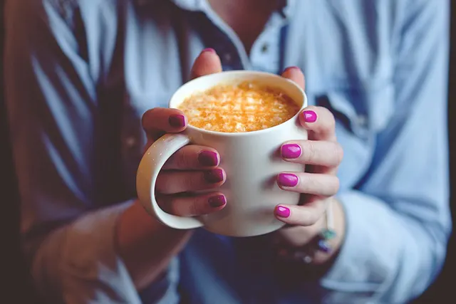 Person in blue shirt holding white mug of hot drink with pink nail varnish.