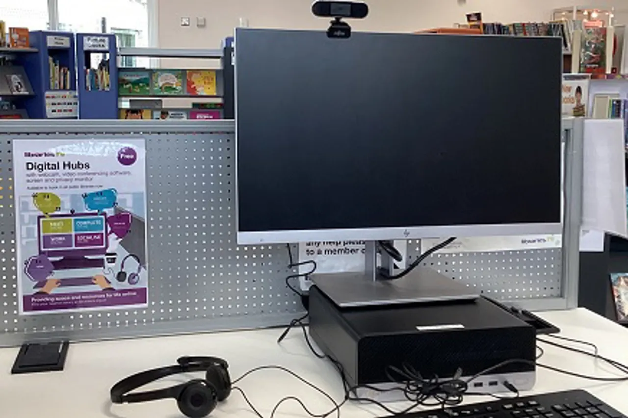 A digital hub workstation with a blank monitor, headphones, and equipment in a library setting surrounded by shelving and educational displays.