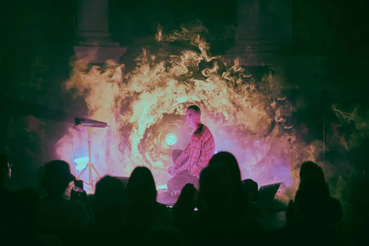 Performer on stage with dramatic fire projection and pink lighting whilst audience members watch from the foreground.