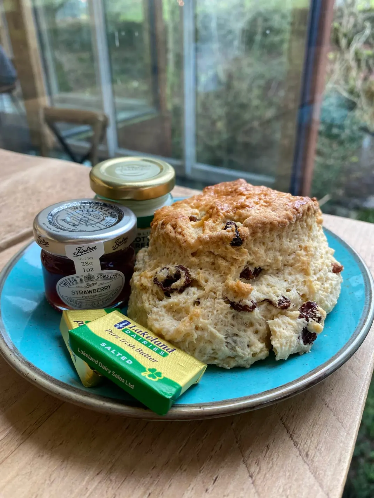 Scone with dried fruit served on a blue plate with butter and strawberry jam, by a garden window.