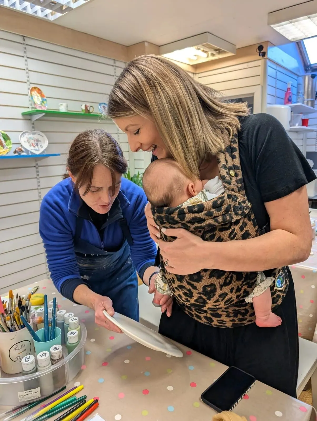 Woman holding a baby and a child in a blue top participate in a ceramic painting activity at a craft table.