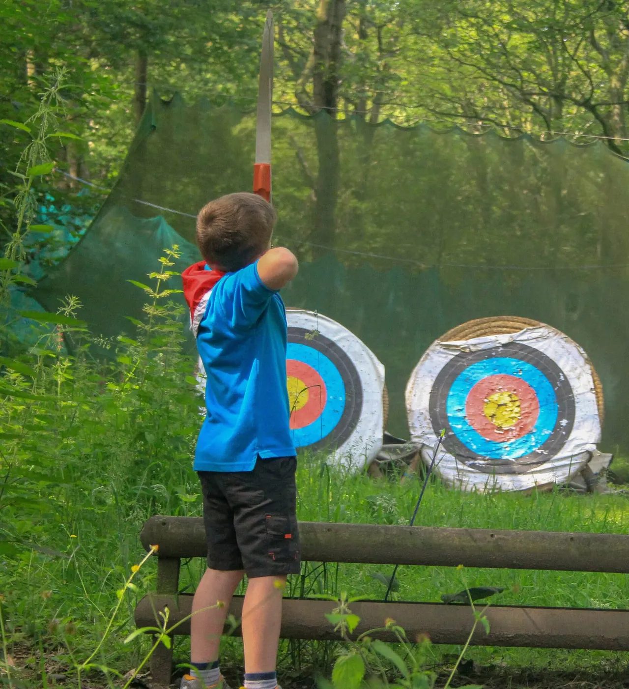 Child in blue jacket aiming at painted target boards in a wooded outdoor activity area.