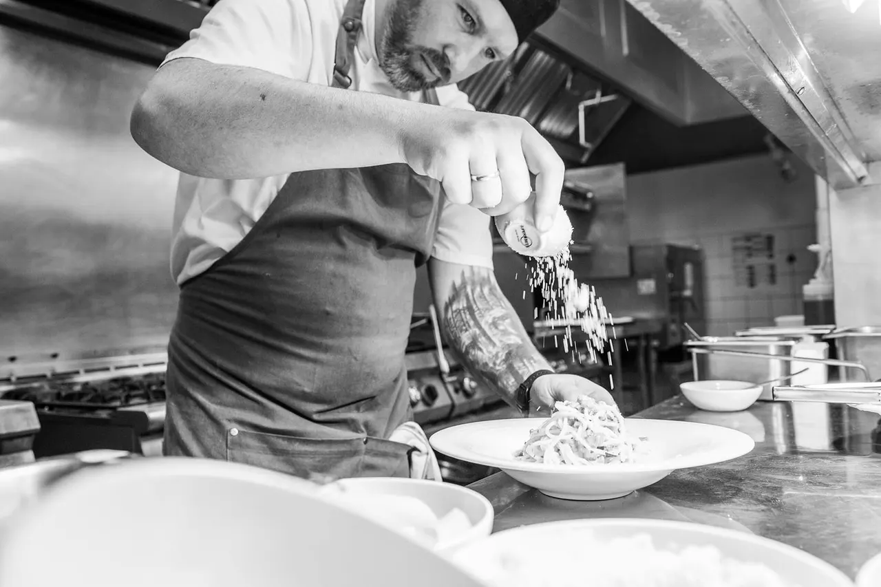 Chef sprinkling grated ingredient onto a plate of pasta in a professional kitchen.