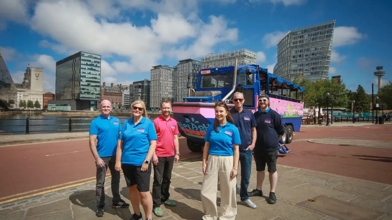 Group of staff members standing in front of a blue and pink amphibious tour bus on a waterfront promenade with city buildings behind.