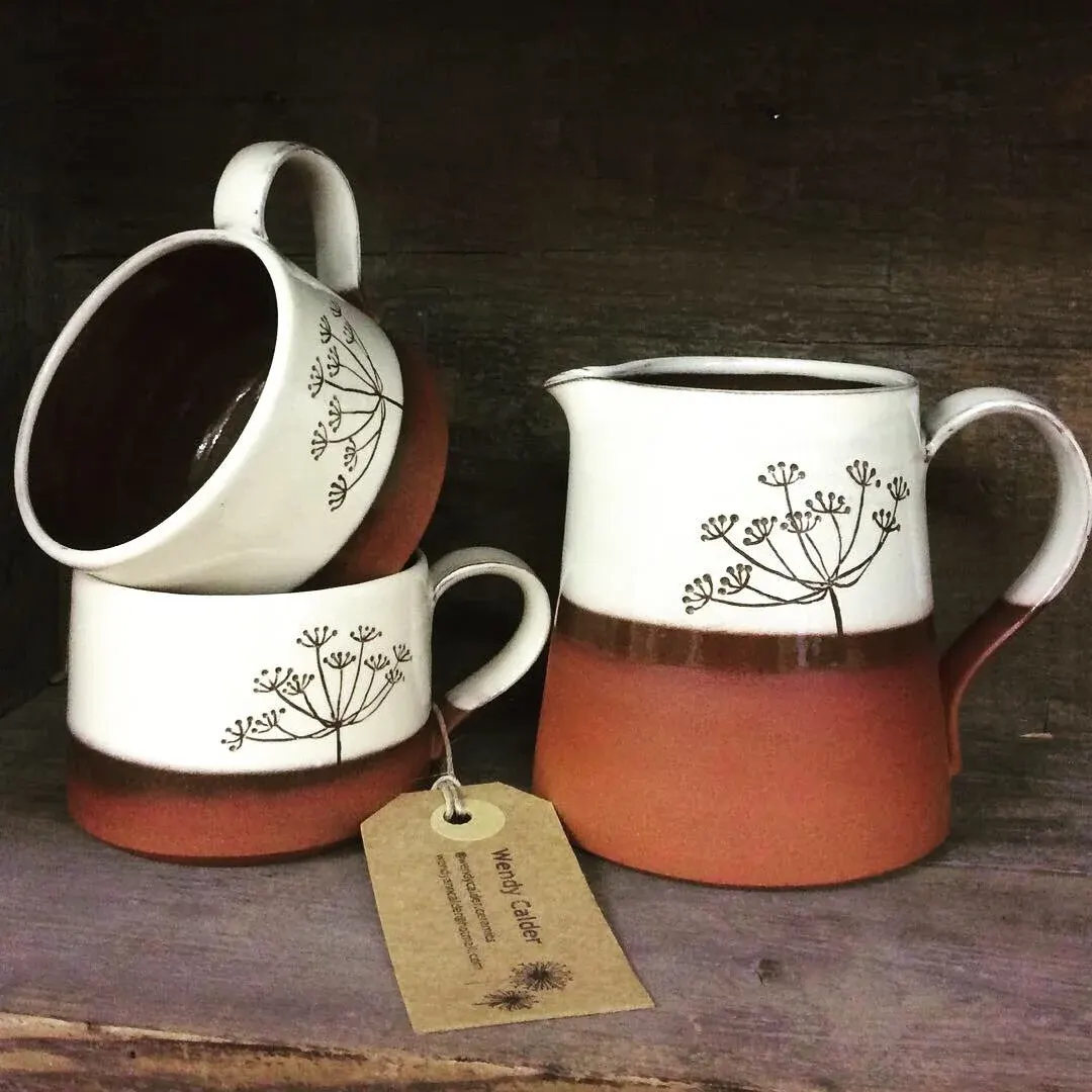 Three ceramic jugs with white upper sections decorated with botanical drawings and terracotta lower sections, displayed on a wooden surface with a maker's tag.