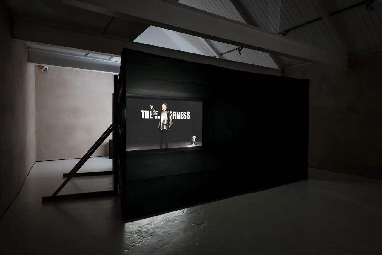 Video installation displayed on a large black screen inside a contemporary gallery space with concrete flooring and skylights.