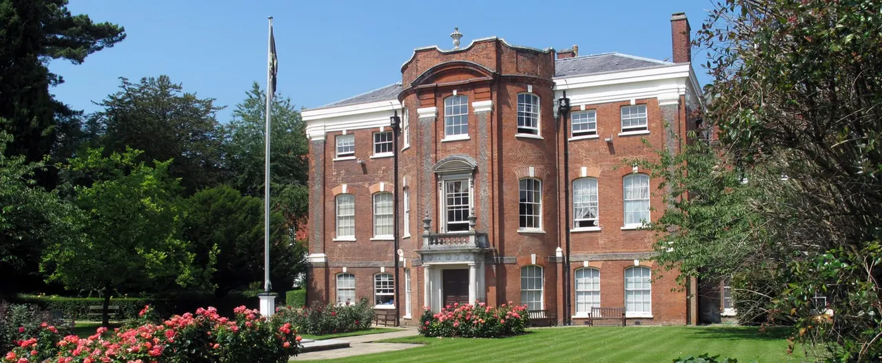 A Georgian brick mansion with manicured lawns and red rose gardens in the foreground, surrounded by mature trees under clear blue sky.