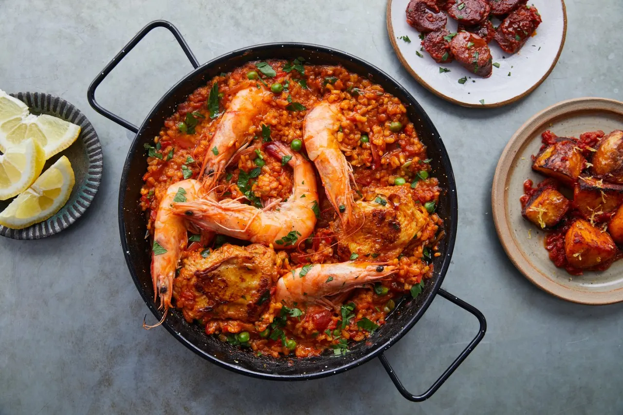 Paella in a large black pan with prawns and saffron rice, surrounded by plates of meat and lemon wedges.