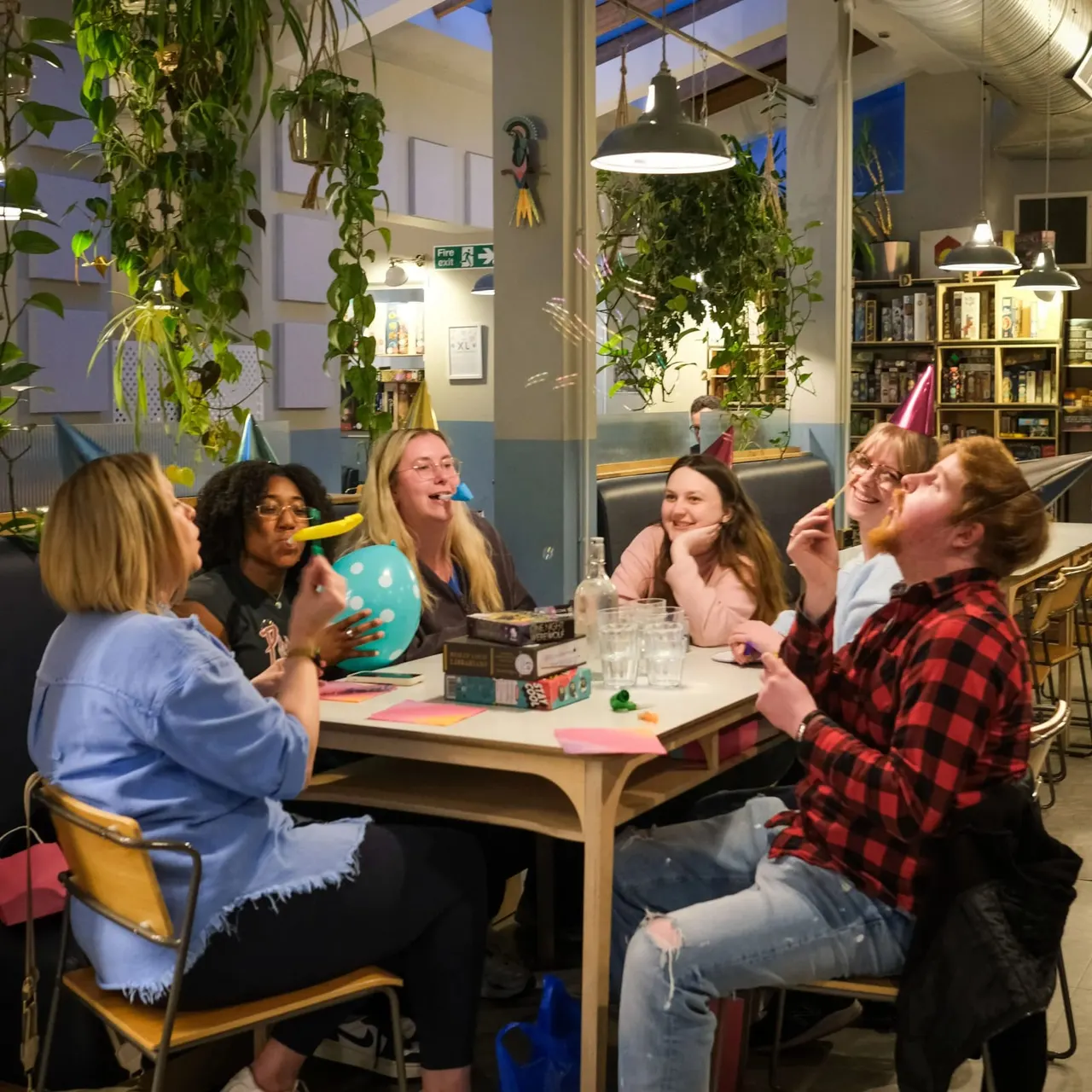 A group of six people wearing party hats gather around a table playing board games and celebrating together in a bright, plant-filled indoor space.