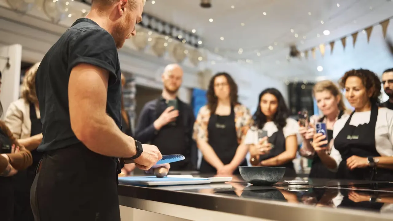 A chef demonstrates cooking techniques to a group of attendees gathered around a preparation counter in a bright, decorated hall.