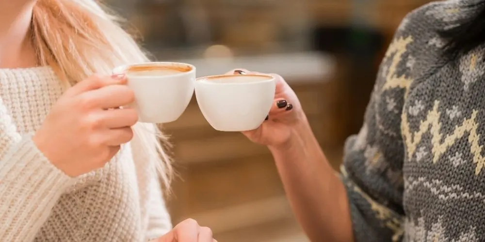 Two people clinking white cups of coffee or hot chocolate together outdoors.