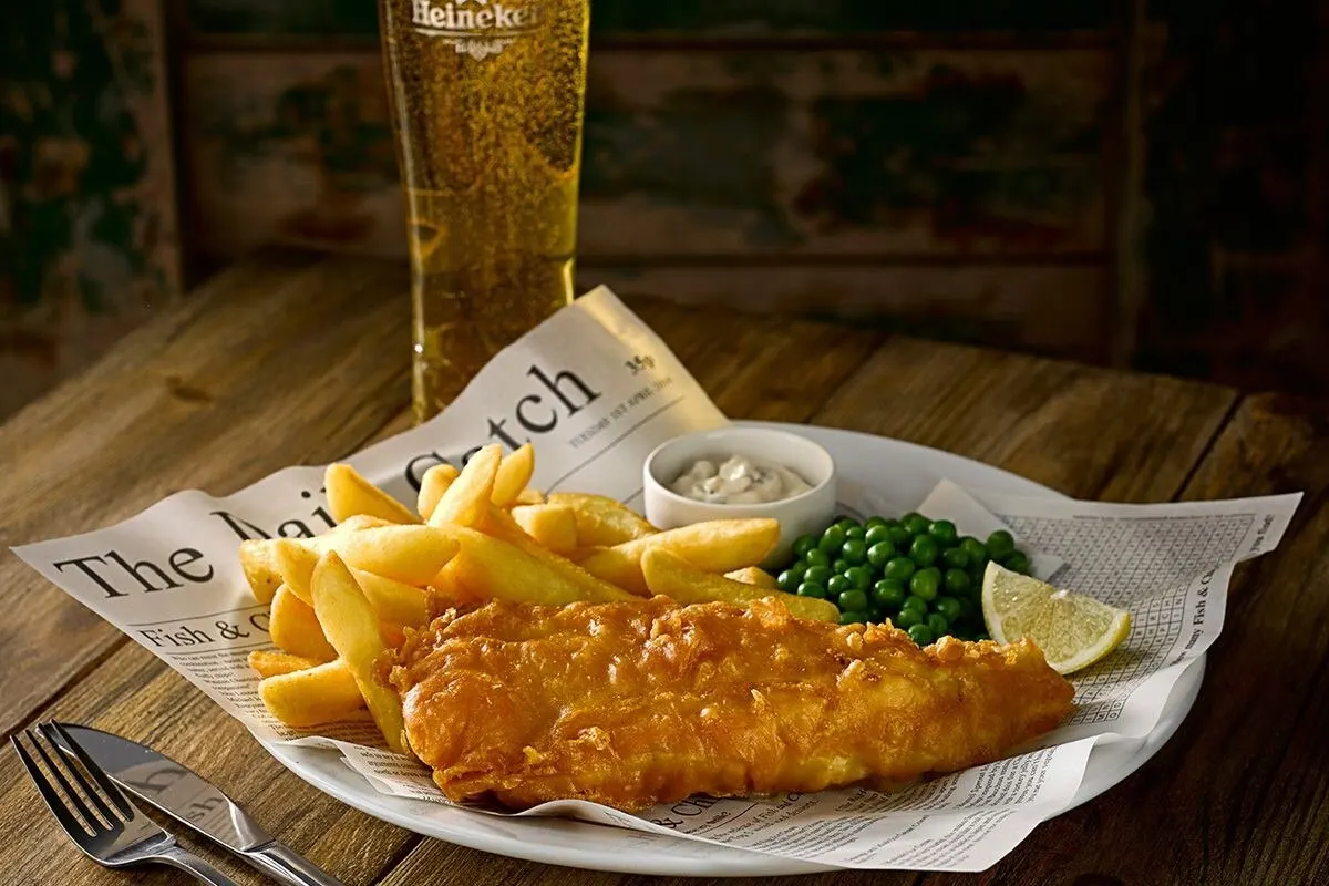 Battered fish fillet with chips, peas, tartare sauce and lemon on newspaper-lined paper, with a glass of lager on a wooden table.