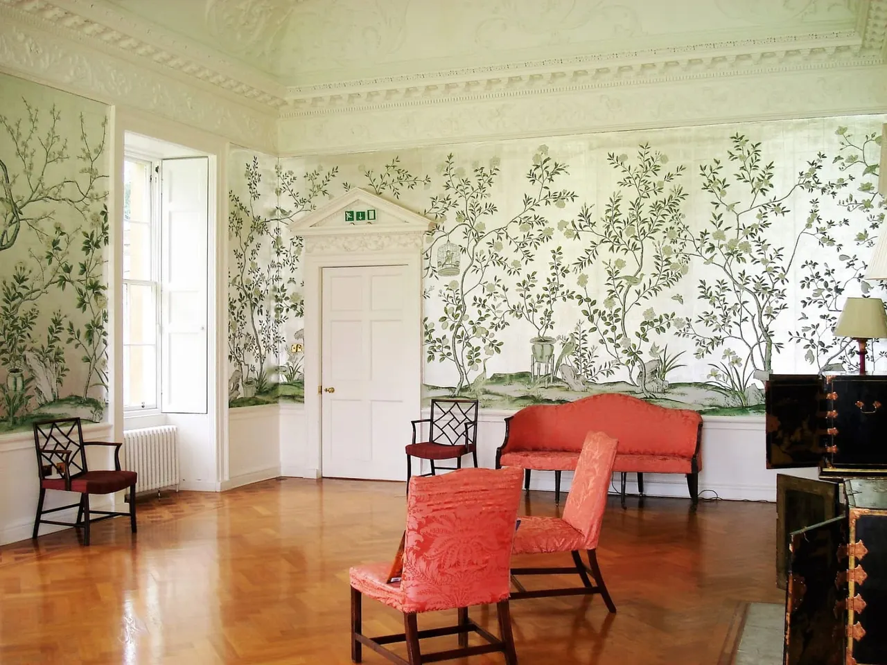 Elegant drawing room with ornate plasterwork ceiling, botanical wallpaper, red upholstered furniture, and polished wooden floors.