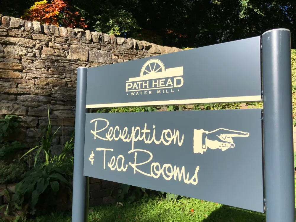 Directional sign for Path Head Water Mill reception and tea rooms, with stone wall and trees behind.