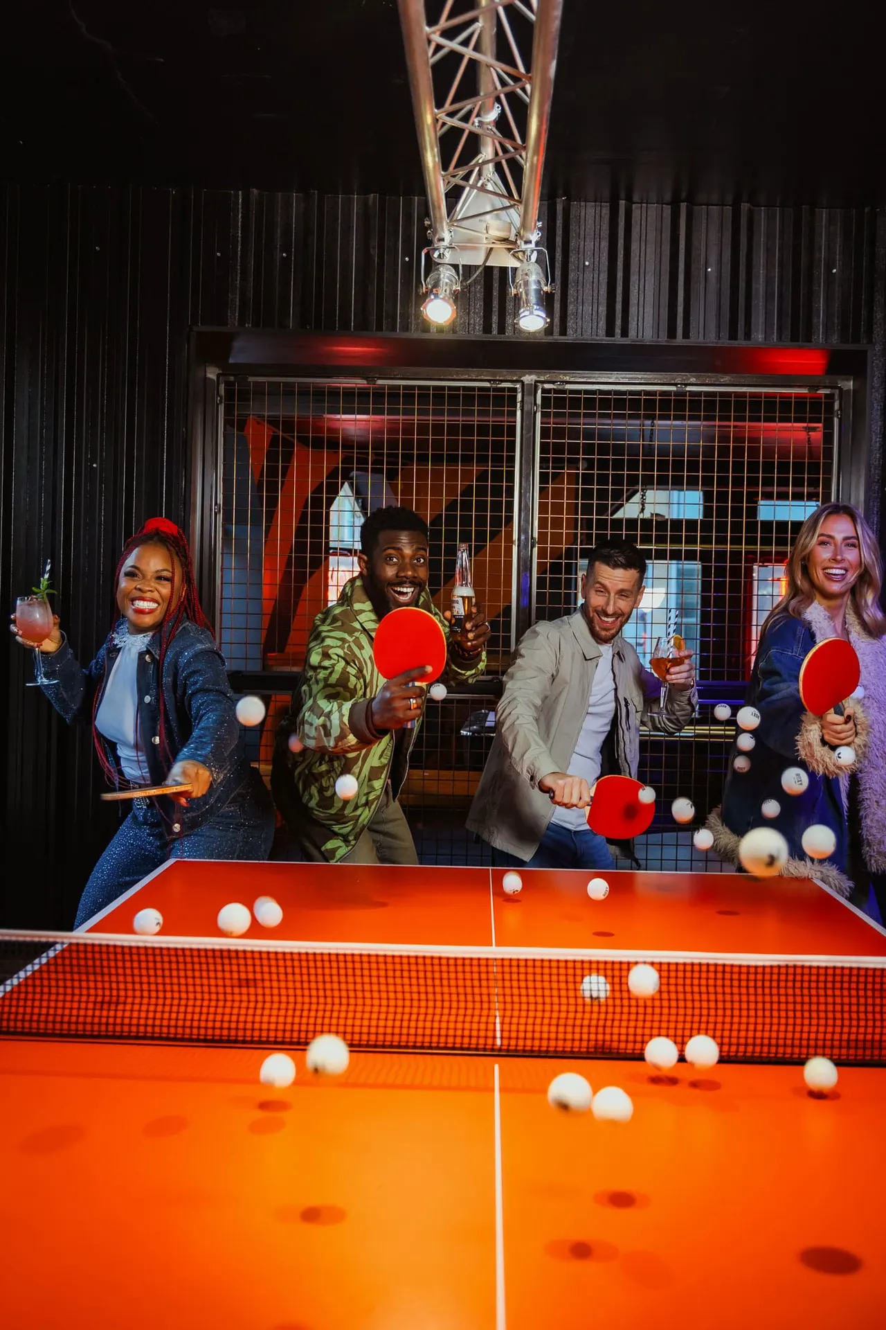 Five people playing table tennis indoors at night, holding red paddles and drinks.