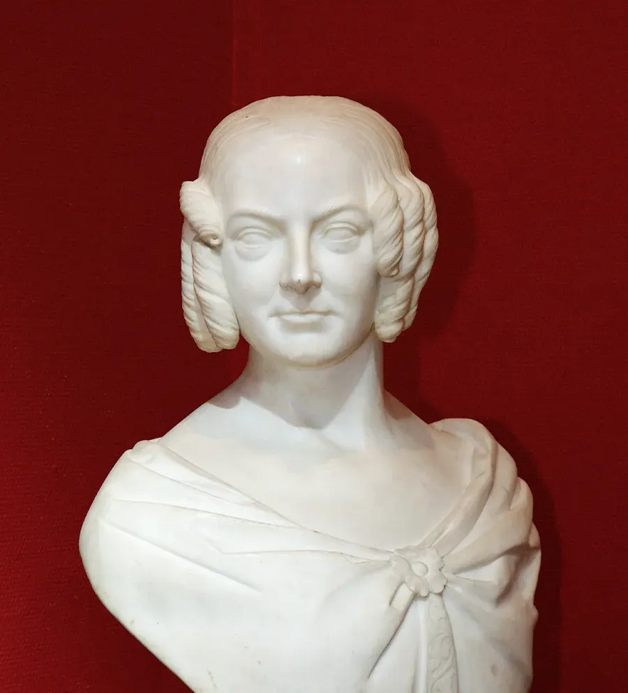 White marble bust of a woman with ringlets on either side of her face, wearing draped clothing, displayed against a deep red wall.