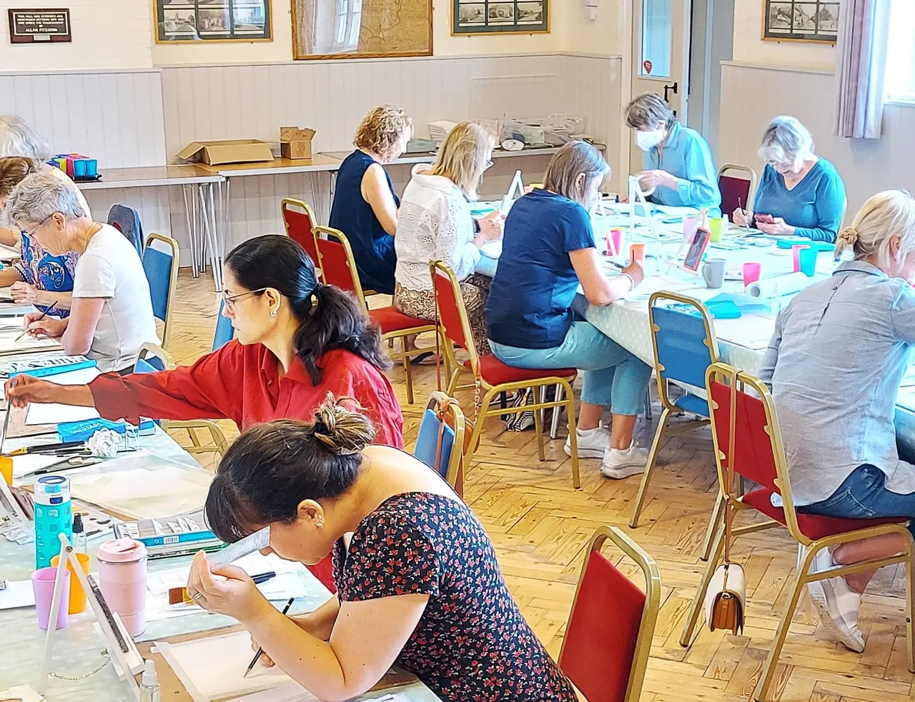 Group of adults seated at tables participating in an art class, with materials and artwork spread across the surfaces.